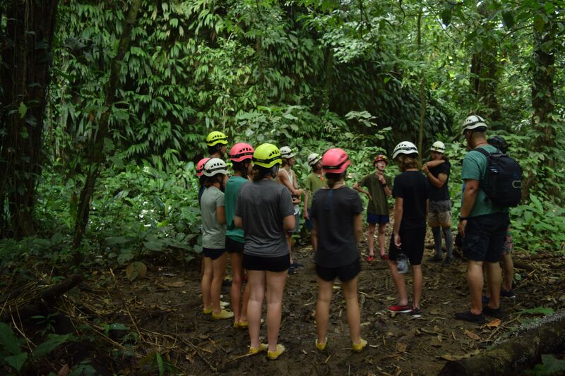 In a lush green forest, a group of people wearing helmets gathers. They appear to be preparing for an outdoor activity, possibly a hike or a guided tour. The dense foliage suggests a tropical or subtropical environment. The group is standing on a muddy ground, indicating recent rain or high humidity.